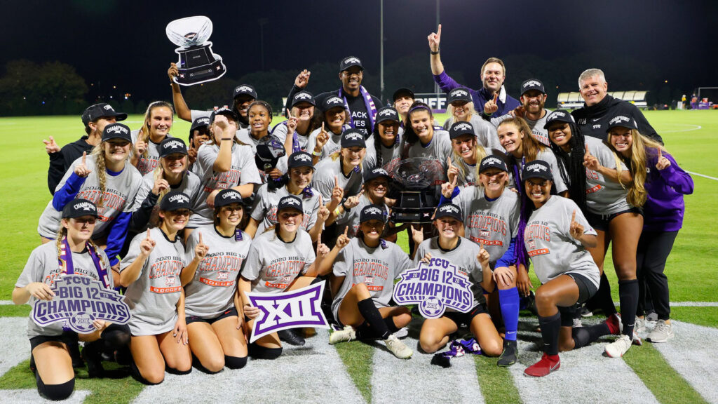 The TCU soccer players and coaches crowds together around the Big 12 conference trophy. The players are all smiling while multipe players hold up cardboard cutouts and the majority hold up a number 1 hand signal.